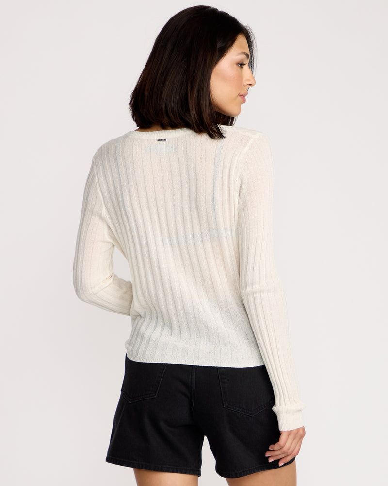 A woman with medium-length dark hair wears the Volcom Women's Pulp Stitchin Cardi, a body-skimming, long-sleeve sheer ribbed sweater, paired with black shorts as she stands facing away from the camera against a white background.