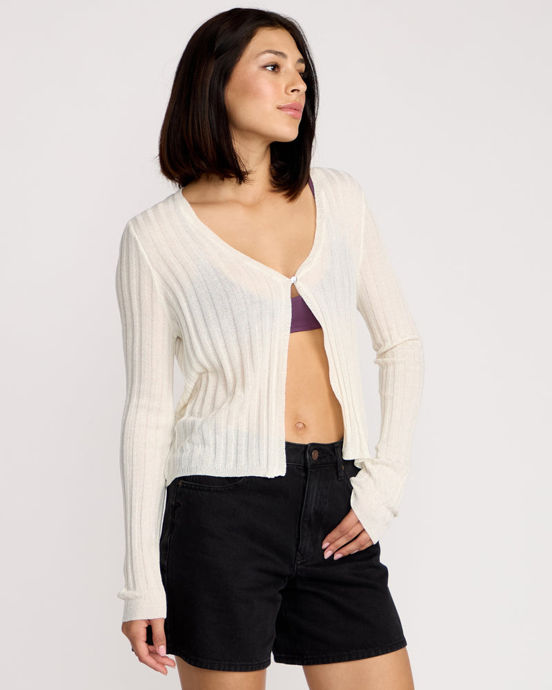 A woman with straight, shoulder-length brown hair wears the Volcom Women's Pulp Stitchin Cardi, cream-colored with fitted sleeves, fastened over a purple bralette and black high-waisted shorts. She stands against a plain white background, looking to the side.