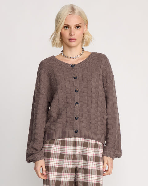 A woman with short blonde hair wears the Volcom Women's Snug Lyfe Cardigan, featuring a relaxed fit and basket-weave knit, paired with plaid pants, standing against a plain white background.