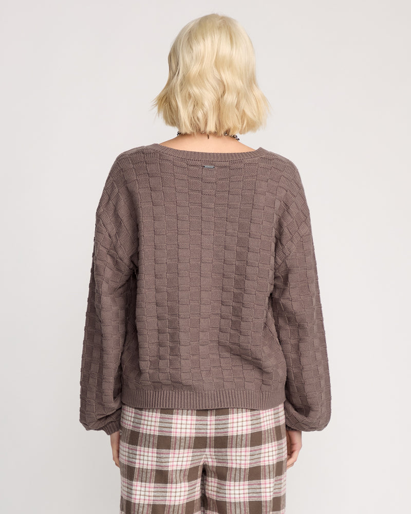 A woman with short, wavy blonde hair is shown from behind in a relaxed fit mauve Volcom Women's Snug Lyfe Cardigan and brown, pink, and white plaid pants, set against a plain light background.