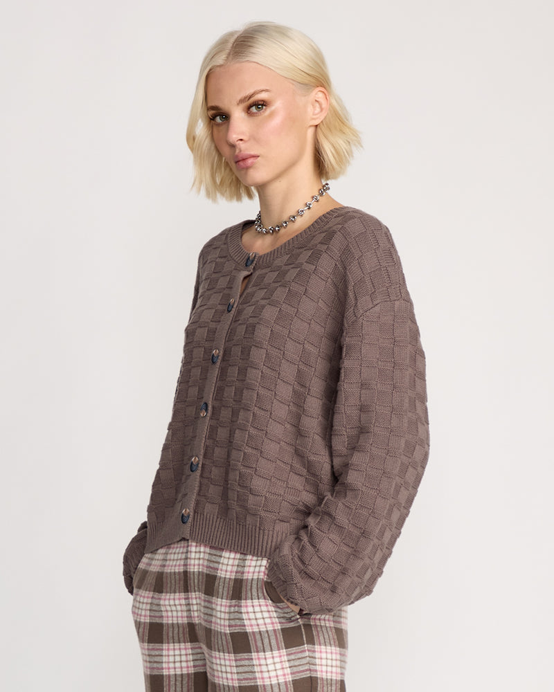 A woman with short blonde hair wears the Volcom Women's Snug Lyfe Cardigan, a relaxed fit brown basket-weave knit button-up, paired with plaid pants. She stands with hands in her pockets against a plain light background.