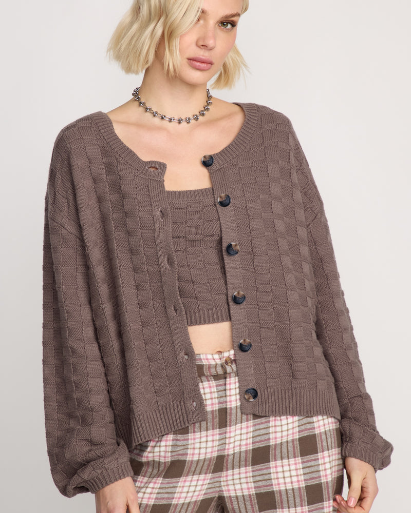 A woman with short blonde hair wears the Volcom Women's Snug Lyfe Cardigan over a matching crop top and plaid pants in brown, pink, and white, paired with a beaded choker necklace.
