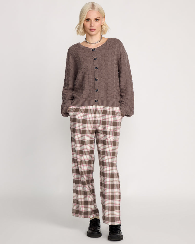 A woman with short blonde hair wears a relaxed fit Volcom Women's Snug Lyfe Cardigan in a brown basket-weave knit, plaid brown-and-white pants, and black shoes, standing against a plain light background.