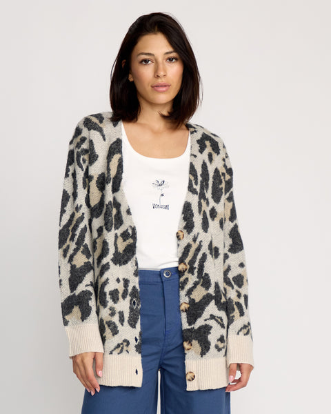 A woman with shoulder-length dark hair wears a white top, blue pants, and the Volcom Women's Wylding Out Cardi—an oversized animal jacquard cardigan with large buttons—standing against a plain light background, facing the camera.