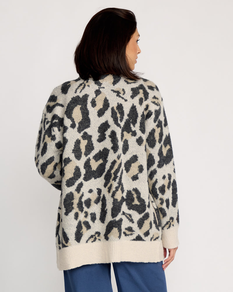 A woman with shoulder-length dark hair faces away, wearing the Volcom Women's Wylding Out Cardi in a cream and black animal jacquard pattern over blue pants, standing against a plain white background.