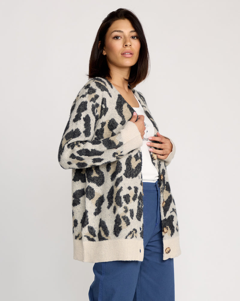 A woman with shoulder-length dark hair wears the Volcom Women's Wylding Out Cardi over a white shirt and blue pants, standing against a plain light background and looking confidently at the camera.