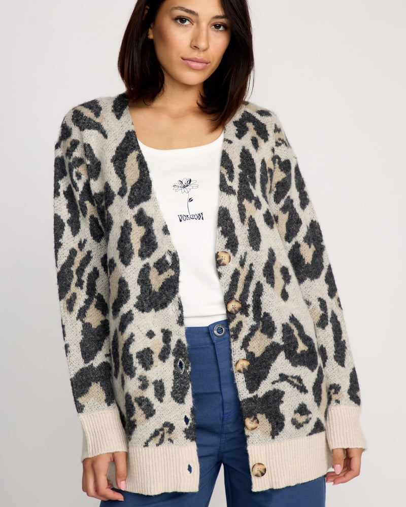 A woman wears the Volcom Women's Wylding Out Cardi—an oversized beige and black animal jacquard cardigan with large buttons—over a white graphic shirt and blue pants, standing against a plain background.