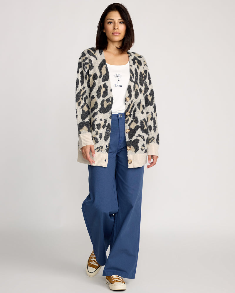 A woman with straight dark hair wears the Volcom Women's Wylding Out Cardi, a white jacquard mohair-wool cardigan with oversized tortoiseshell buttons, paired with a white shirt, blue wide-leg pants, and tan sneakers against a plain background.