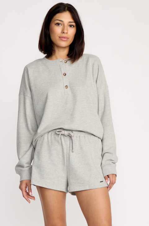 A woman poses against a plain background wearing Volcom Women's Lived In Fleece Shorts with an elastic waistband, paired with a light gray, long-sleeve button-up pullover, looking calmly at the camera.