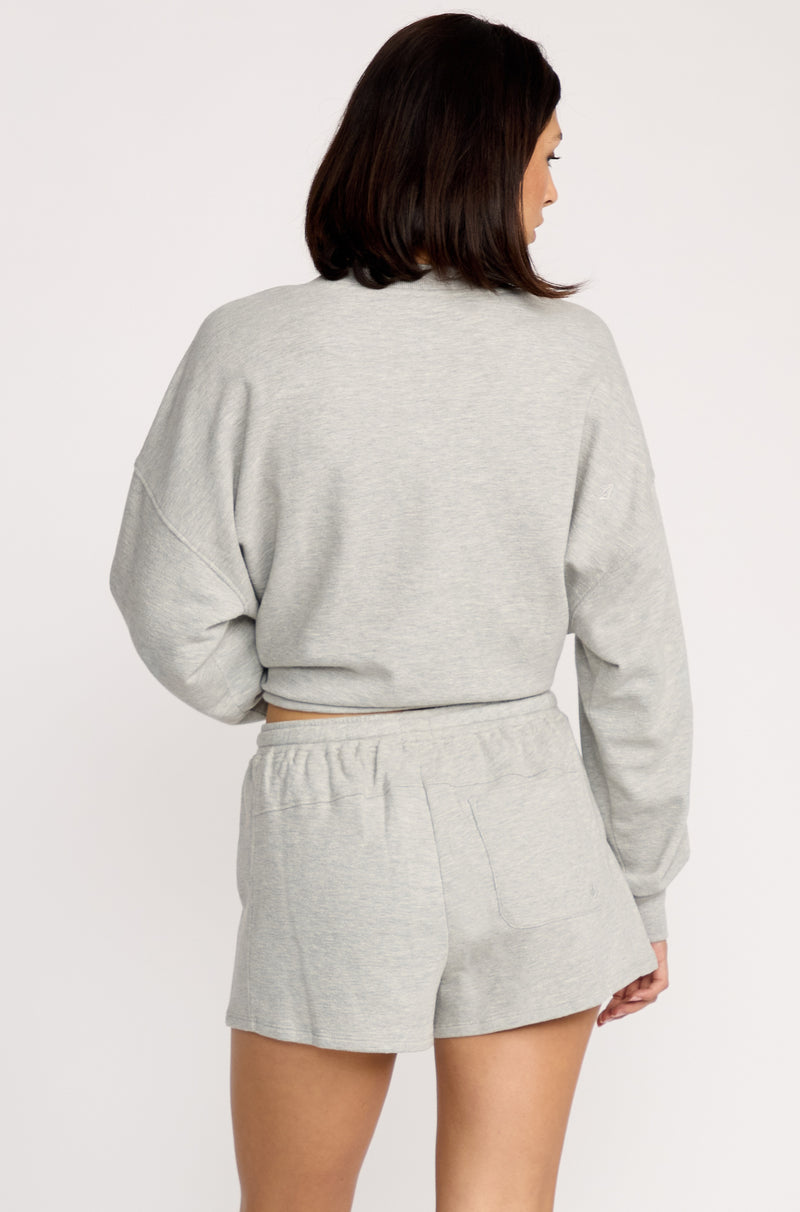 A woman with shoulder-length brown hair, seen from behind, wears a light gray long-sleeve sweatshirt and Volcom Women's Lived In Fleece Shorts with an elastic waistband. The background is plain white.