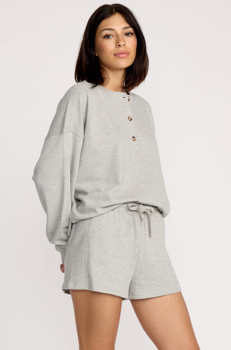 A woman with shoulder-length dark hair wears Volcom Women's Lived In Fleece Shorts, featuring an elastic waistband, paired with a loose-fitting, light grey long-sleeve henley shirt. She stands against a plain light background.