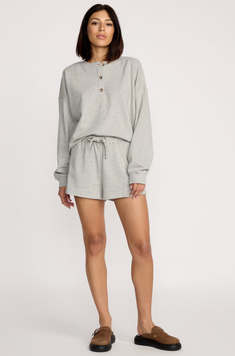 A woman with shoulder-length dark hair wears brown slip-ons and Volcom Women's Lived In Fleece Shorts with an elastic waistband, paired with a light gray long-sleeve henley top, standing against a plain background.