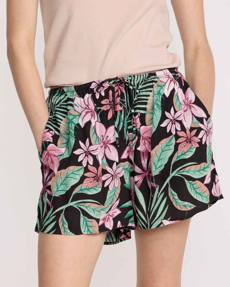 A person wears Volcom Women's Boss Beach Shorts featuring a vibrant pink and green tropical floral print, paired with a light pink top and hands in pockets.