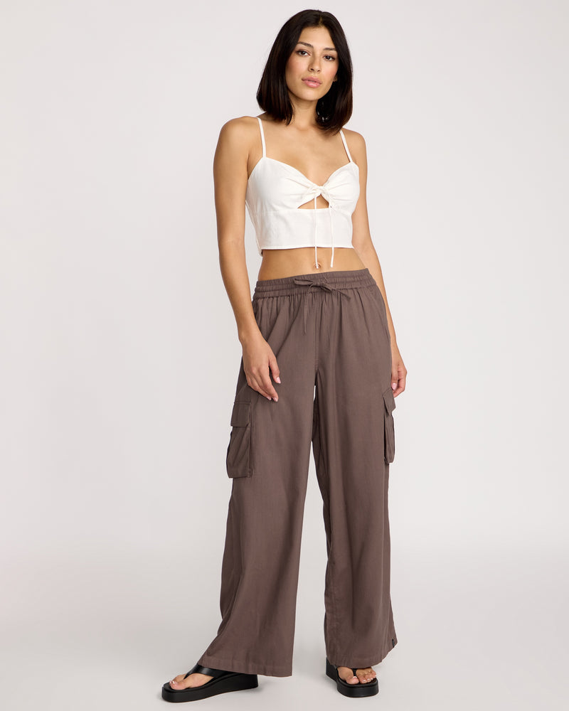 A woman with straight, shoulder-length dark hair stands against a plain background wearing Volcom Women's Lived In Beach Pants in brown, paired with a white cropped top with thin straps and front tie detail, and black sandals.