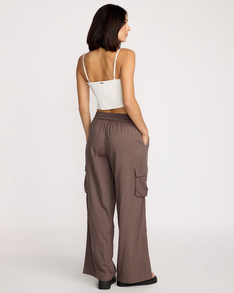 A woman with shoulder-length dark hair faces away, wearing Volcom Women's Lived In Beach Pants in brown with cargo pockets and a white cropped tank top, standing against a plain light background.