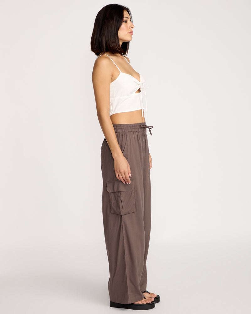 A woman stands in profile wearing a white crop top, Volcom Women's Lived In Beach Pants in brown with cargo pockets, and black sandals against a plain light background.