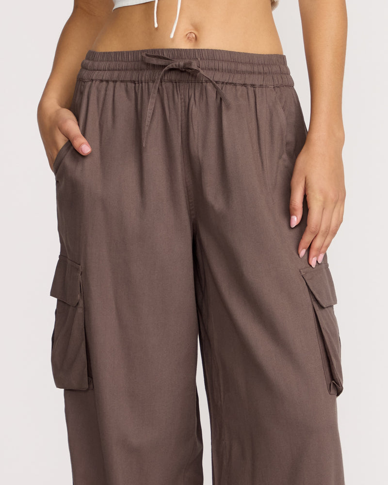 A person wears Volcom Women's Lived In Beach Pants—loose-fit brown cargos with cargo pockets and a drawstring waistband—standing with one hand in a pocket and the pant's cropped top plus their midsection visible.