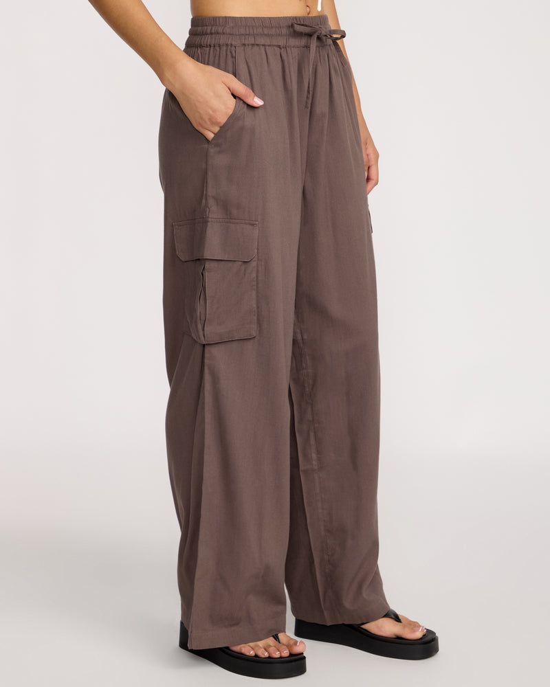 A person wears Volcom Women's Lived In Beach Pants in brown with cargo pockets and black sandals, standing with one hand in a pocket against a plain light background.
