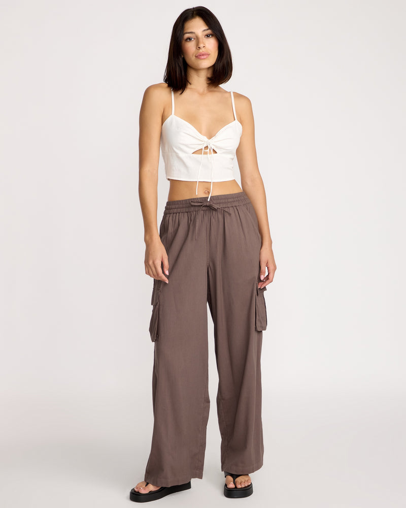 A woman with shoulder-length dark hair and a neutral expression models Volcom Women's Lived In Beach Pants, featuring cargo pockets, with a white strappy crop top and black sandals against a plain background.