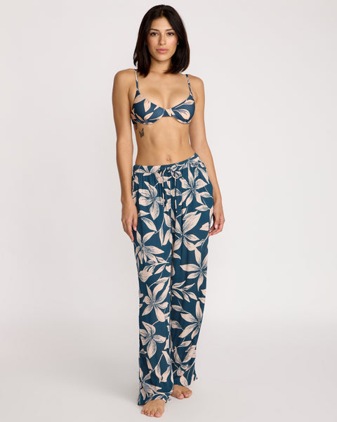 A woman stands barefoot in Volcom Women's Shore Shot Pants in Harbor Blue and a bralette with a light leaf pattern, set against a plain light background.