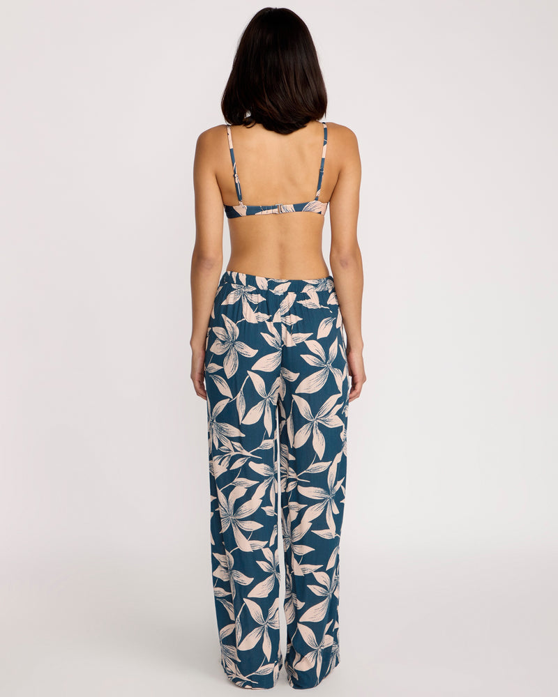 A woman faces away, wearing a strappy bikini top and Volcom Women's Shore Shot Pants in Harbor Blue with a blue and white leaf pattern. Her arms rest at her sides against a plain white background, showcasing chic beachwear style.