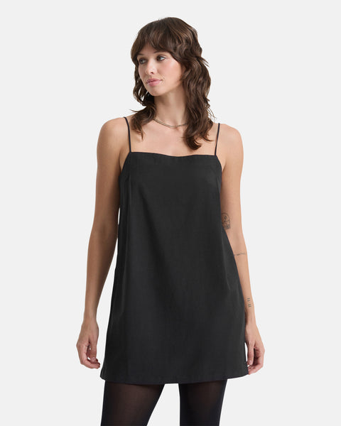 A woman with wavy, shoulder-length brown hair wears the Women's Backbeatz Dress by Volcom—a sleeveless black dress with thin straps—paired with black tights, standing against a plain white background.