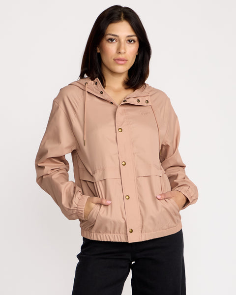 A woman with medium-length dark hair wears the Volcom Women's Stonewind Jacket in light pink, featuring a hooded zip front, snap buttons, front pockets, and a coated taffeta shell, paired with black pants against a plain white background.