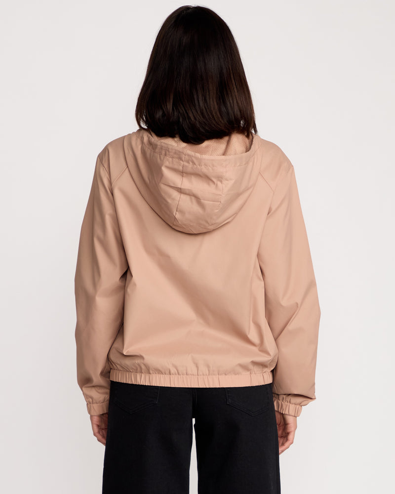 A woman with shoulder-length dark hair is seen from the back wearing a Volcom Women's Stonewind Jacket in beige, featuring a hood and zip front, paired with black pants, standing against a plain white background.