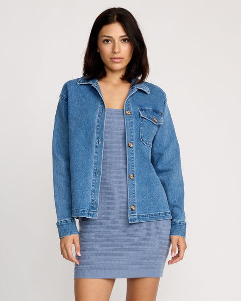 A woman with shoulder-length dark hair and a neutral expression wears the Volcom Women's Stoned BF Shacket in denim over a light blue textured sleeveless dress, standing against a plain background.