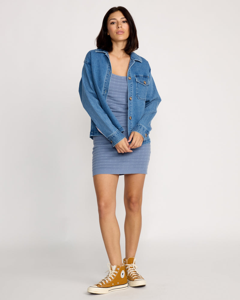 A woman with shoulder-length dark hair stands confidently against a plain backdrop wearing the Volcom Women's Stoned BF Shacket over a fitted blue dress and mustard yellow high-top sneakers.