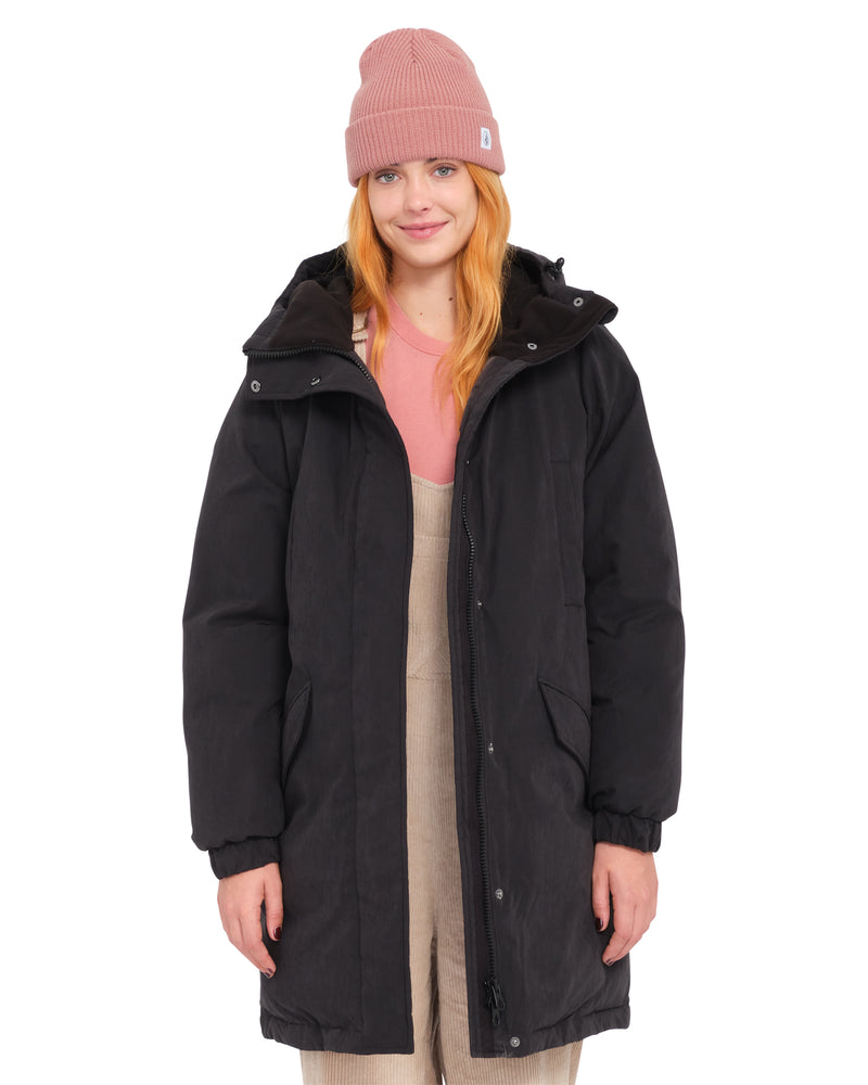 A person wearing a Volcom Women’s Sleepi Puff Up Parka over a pink shirt and tan overalls, topped with a pink knit beanie.