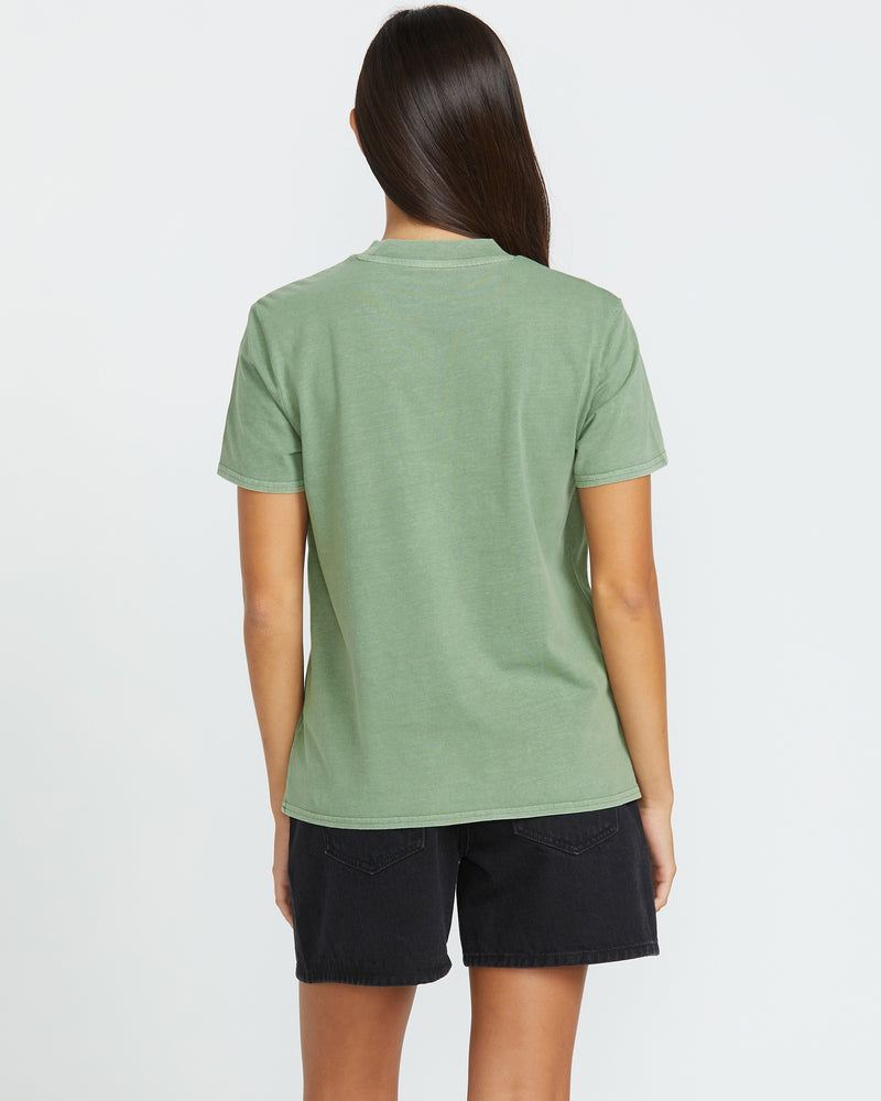 A woman with long dark hair, seen from behind, wears the Volcom Women's Lock It Up Tee in light green with black denim shorts. Her relaxed style stands out against the plain white background.