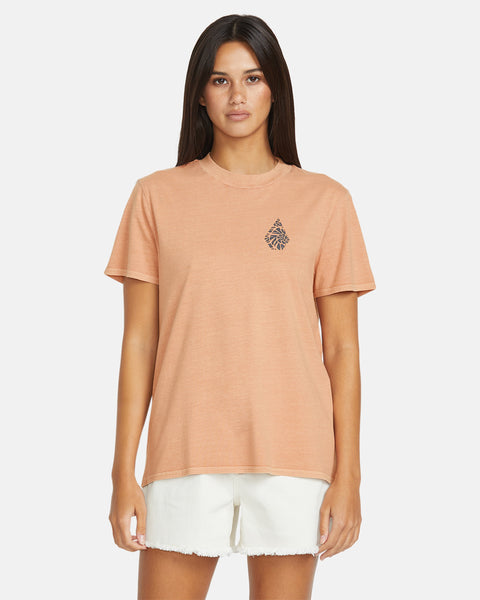 A woman with long dark hair wears the Volcom Women's Lock It Up Tee, an organic cotton peach-colored shirt with a small black chest graphic, paired with white shorts, standing against a plain white background.