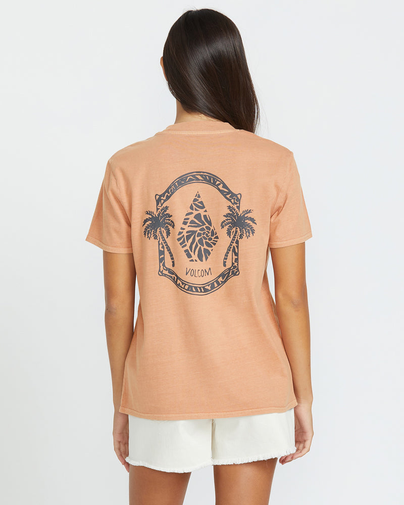 A woman with long brown hair wears the Volcom Women's Lock It Up Tee, a peach-colored organic cotton shirt featuring a black leaf, two palm trees, and VOLCOM text graphic on the back, paired with white shorts against a plain backdrop.