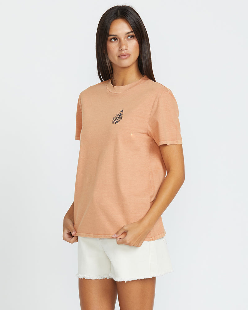 A woman with long dark hair wears the Volcom Women's Lock It Up Tee in peach with a small black leaf design on the chest, paired with white shorts, standing against a plain white background.