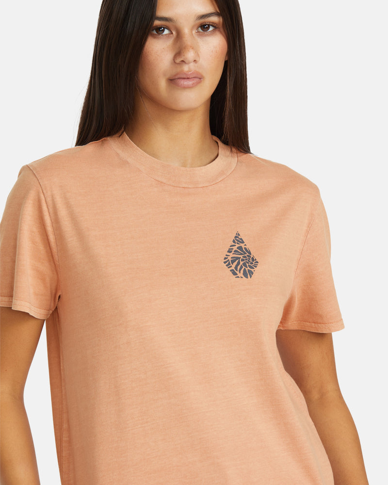 A woman with straight dark hair wears the Volcom Women's Lock It Up Tee, an organic cotton t-shirt in peach with a small dark leaf design on the left chest. She stands against a plain white background, looking forward neutrally.