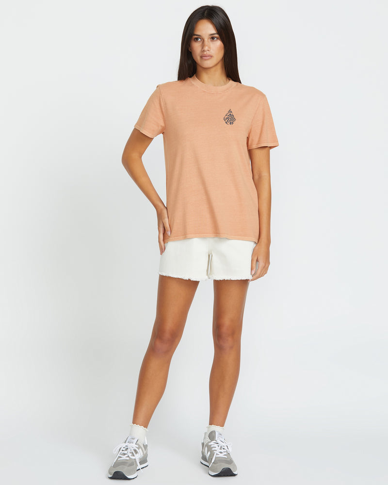 A woman faces forward against a plain white background wearing the Volcom Women's Lock It Up Tee in peach with a small chest graphic, paired with white frayed-hem shorts, white socks, and gray sneakers.