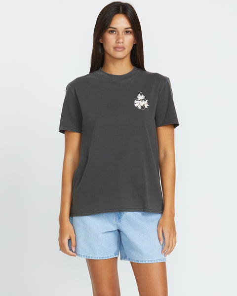 A woman with long dark hair wears the Volcom Women's Lock It Up Tee in dark gray, featuring a small left chest graphic, paired with light blue denim shorts and standing against a plain white background.