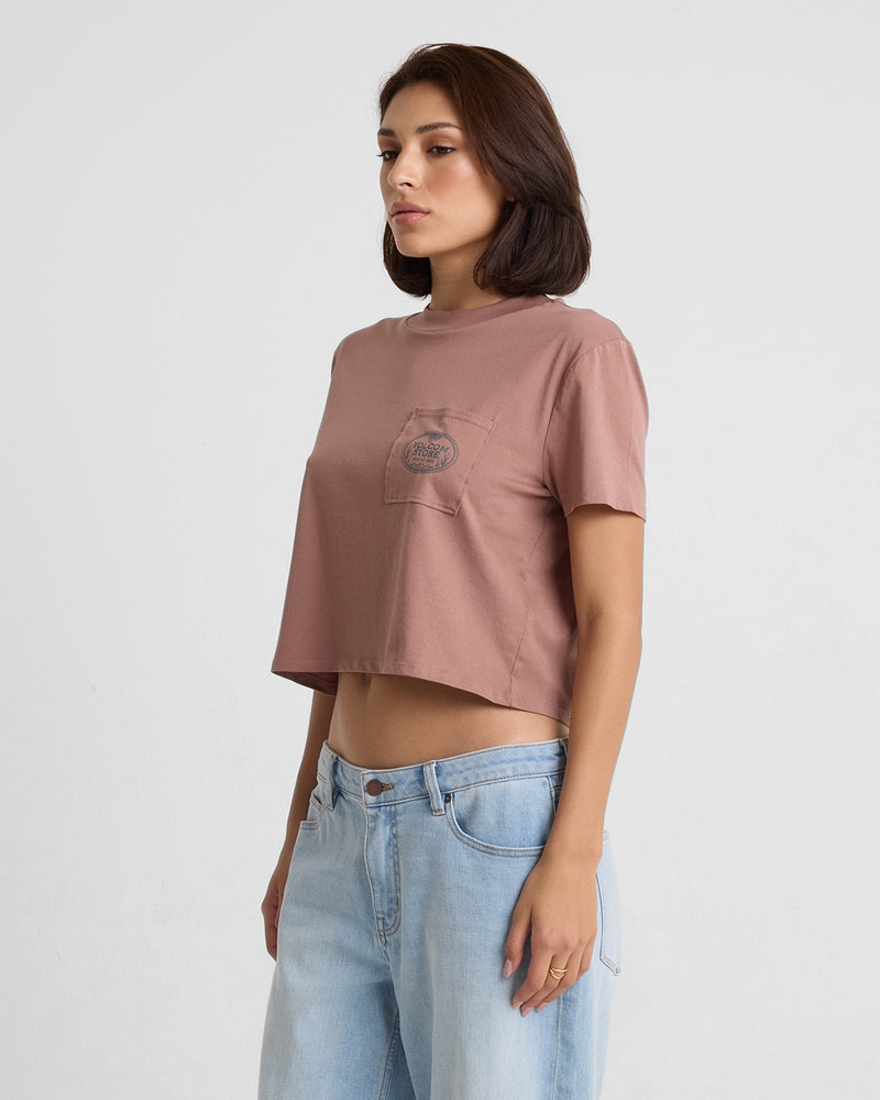 A woman with shoulder-length brown hair and a neutral expression stands against a plain background, wearing a mauve Volcom Women's Pocket Dial Short Sleeve Tee with a small pocket and light blue jeans, looking slightly to the side.