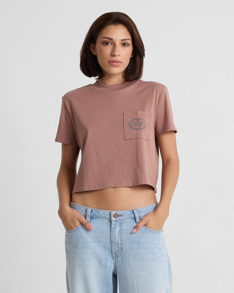 A woman with short brown hair wears a Volcom Women's Pocket Dial Short Sleeve Tee in mauve and light blue jeans, standing with her hands in her pockets against a plain white background.