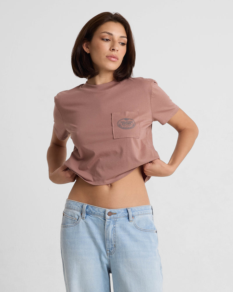 A woman wearing the Volcom Women's Pocket Dial Short Sleeve Tee in mauve and light blue jeans stands against a plain white background, slightly lifting her shirt and looking to the side.