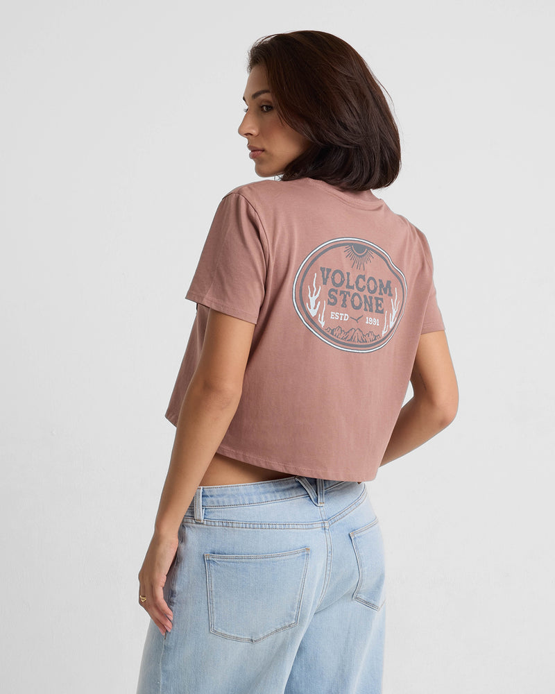 A woman with shoulder-length brown hair wears the Volcom Women's Pocket Dial Short Sleeve Tee, featuring a circular VOLCOM STONE ESTD 1991 back graphic. She pairs it with light blue jeans and faces away in a minimal studio setting.