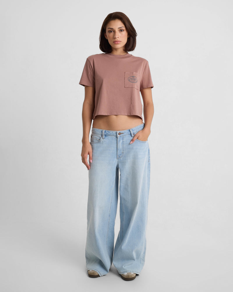 A woman stands against a white background wearing the Volcom Women's Pocket Dial Short Sleeve Tee and loose, light blue wide-leg jeans, with one hand in her pocket and a neutral expression.