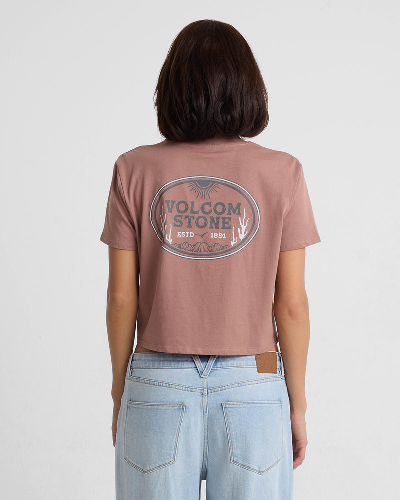 A person with shoulder-length dark hair, wearing the Volcom Women's Pocket Dial Short Sleeve Tee in brown and light blue jeans, stands facing away from the camera against a plain white background.