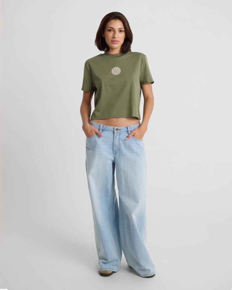 A woman stands against a plain white background, wearing the Volcom Women's Dial Crop Tee in olive green with a white sun graphic and high-waisted, wide-leg light blue jeans, hands in her pockets.