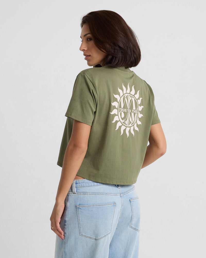 A woman with short dark hair wears Volcom’s Women’s Dial Crop Tee in olive green organic cotton, featuring a white sun graphic and text on the back, paired with light blue high-waisted jeans against a plain light background.