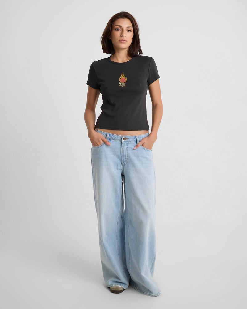 A woman with straight brown hair poses against a white background, wearing the Volcom Women's Have A Clue Tee—a fitted black t-shirt—and light blue, wide-leg jeans, with her hands in her pockets.
