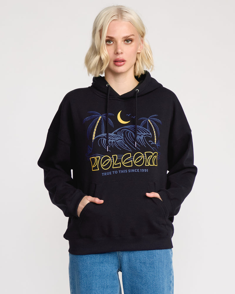 A woman with blonde hair faces forward, hands in her pocket, wearing the Volcom Women's Stone Art Up Hoodie in black with palm trees, waves, and moon graphics and yellow text. Its brushed fleece and relaxed fit pair well with her blue jeans.