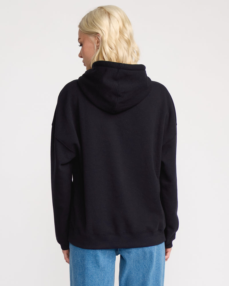 A blonde woman wearing the Volcom Women's Stone Art Up Hoodie with a dropped-shoulder fit and blue jeans stands with her back to the camera against a plain white background.