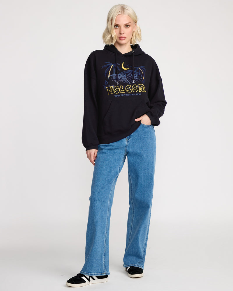A woman with short blonde hair wears a black Volcom Women's Stone Art Up Hoodie with a dropped-shoulder fit, blue jeans, and black sneakers, standing against a plain white background.